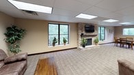 This single-floor space is thoughtfully designed to provide a comfortable and functional environment for gathering and relaxation. The interior is finished with warm beige walls, neutral plaid carpeting, and a drop tile ceiling with recessed panel lighting throughout, creating a consistently inviting atmosphere across all areas.

The main lounge area is a standout feature, centered around a stacked-stone fireplace with a dark mantle and a wall-mounted flat-screen television above it. Large windows line the walls, flooding the space with natural light and offering pleasant views of surrounding greenery. Potted plants accent the room, adding a touch of warmth and life.

Adjacent to the lounge is a comfortable seating room furnished with plush, oversized sofas and a rustic wooden coffee table, complemented by decorative wall art. This area is well-suited for casual conversation or relaxation.

The dining area accommodates multiple groups simultaneously, featuring sturdy wooden tables and matching slat-back chairs arranged on the carpeted floor. A row of windows continues to bring in natural light and outdoor views of the tree line.

The kitchen is fully equipped with dark espresso cabinetry, light stone-look countertops, stainless steel appliances including a refrigerator, electric range, and over-the-range microwave, making it highly functional for food preparation and serving.