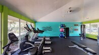 This fitness center is a well-appointed workout facility designed to support a full range of exercise routines. The space is immediately distinguished by its bold color scheme, featuring vivid lime green and teal accent walls adorned with an inspirational fitness-themed word art mural. The vaulted ceiling adds a sense of openness, while recessed lighting and a ceiling fan contribute to a comfortable workout environment.

Large windows line multiple walls, flooding the space with natural light and offering views of the surrounding outdoor greenery and pool area. Full-length mirrors along one wall enhance the sense of depth and allow users to monitor their form during exercise.

The cardio zone is equipped with treadmills, elliptical machines, a stationary bike, and a rowing machine, all positioned to take advantage of the outdoor views. The strength training area includes a multi-station cable machine, a leg extension/curl machine, a weight bench, and a well-stocked dumbbell rack covering a broad range of weights. A dedicated functional fitness corner houses stability balls, medicine balls, and resistance equipment stored on a vertical rack.

The rubber flooring throughout provides cushioning and durability, making the space practical for a variety of workout styles. A wall-mounted television adds convenience for entertainment during workouts.