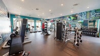 Located at Savona Point in Cape Coral, Florida, this fitness center is a thoughtfully designed, single-floor facility divided into two distinct exercise rooms that together provide a comprehensive workout experience. The main exercise room makes an immediate visual impact with bold, vibrant teal accent walls adorned with motivational health and wellness typography murals, creating an energizing atmosphere. This room features rubber flooring and is generously outfitted with a wide array of cardio equipment, including treadmills, ellipticals, and StairMaster machines, alongside a robust selection of strength training equipment such as cable machines, weight racks, and multi-functional resistance stations. Large windows allow natural light to fill the space while offering views of the surrounding Florida landscape. Recessed lighting provides consistent illumination throughout. The second exercise room offers a calmer aesthetic, with soft sage-green walls, light wood-look flooring, and a clean, open layout that invites functional and group training. This room houses a dedicated cycling area with stationary bikes, a wall-mounted smart display, kettlebell and medicine ball racks, battle ropes, and additional free weights. Together, the two rooms form a complete fitness destination suited for cardio, strength, cycling, and functional training.