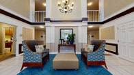Located at 1015 Sibley Memorial Hwy in Saint Paul, this residential community offers a refined and comfortable living environment across its thoughtfully designed common spaces. The grand entrance lobby immediately impresses with its soaring two-story atrium, anchored by an elegant chandelier and framed by upper-level balconies with white railings. Rich blue carpeting, patterned accent chairs, and a console table with lush greenery create a warm and welcoming first impression.

Wide, well-lit corridors with patterned carpeting connect residents to key amenities, including a dedicated Club Room. This expansive gathering space features a coffered ceiling, large windows that flood the room with natural light, and sweeping outdoor views. Comfortable leather seating, patterned armchairs, a wall-mounted television, and multiple dining tables make it an ideal space for both relaxation and socializing.

A fully equipped kitchen with rich hardwood floors, dark cabinetry, stainless steel appliances, granite countertops, and a center island with bar seating adds a functional and elegant touch. Throughout the community, warm wall sconces, neutral tones, and classic furnishings contribute to a consistently inviting and upscale ambiance suited for everyday living.