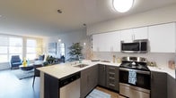 This single-floor residence showcases a thoughtfully designed open-concept layout that blends modern aesthetics with everyday functionality. The kitchen is a standout feature, boasting a two-tone cabinetry scheme with crisp white upper cabinets and charcoal lower cabinets, all complemented by clean white countertops. Stainless steel appliances — including a range, microwave, and dishwasher — lend a polished, contemporary feel. A peninsula with an undermount sink serves as both a practical workspace and a natural divider between the kitchen and living area, with stylish pendant lights hanging above to enhance the ambiance. Warm hardwood-style flooring runs throughout, adding texture and continuity to the space. The kitchen opens directly into the living area, which benefits from the open sightlines and an airy, well-lit atmosphere created by a flush ceiling light and natural light filtering in from nearby windows. The overall design is clean and modern, with neutral tones and carefully chosen décor that create a welcoming, move-in-ready environment. The seamless flow between spaces makes this residence feel expansive and ideal for both everyday living and entertaining.