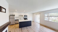 This single-floor residence showcases a clean, modern design with an open-concept layout that seamlessly connects the kitchen and living area. The kitchen is a standout feature, equipped with stainless steel appliances including a refrigerator, range, microwave, and dishwasher. Crisp white cabinetry extends to the ceiling, complemented by a patterned tile backsplash and light quartz countertops. A navy blue center island provides additional prep space and acts as a natural divider between the kitchen and the open living area. Light hardwood-style flooring runs throughout, lending warmth and continuity to the space. The living area is bathed in natural light, courtesy of a large window and sliding glass French doors that open to a balcony, offering outdoor views and ventilation. Recessed lighting throughout ensures bright, even illumination. A fireplace with built-in shelving anchors one wall of the living space, adding character and a focal point to the room. The bathroom is finished with a clean, minimalist aesthetic, featuring a white vanity with an undermount sink, a framed mirror, and wood-tone flooring. The overall ambiance is bright, airy, and contemporary, reflecting a thoughtfully designed living environment.