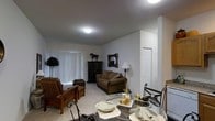 This single-floor residence presents an open-concept layout that seamlessly integrates the living and dining areas into one cohesive space. The living area is anchored by a brown upholstered sofa accented with decorative pillows, complemented by wooden armchairs that add a warm, traditional character. A dark wood cabinet serves as a focal point along the wall, topped with a decorative horse figurine, while a rustic framed mirror adds visual interest above. A floor lamp provides supplementary ambient lighting alongside the flush-mounted ceiling fixtures that illuminate the space evenly.

The dining area features a glass-top round table surrounded by wrought iron chairs, elegantly set with folded linens and decorative accessories including candles and gold-toned glassware. The light-toned flooring throughout creates a sense of continuity between the two areas.

Vertical blinds on the windows allow for adjustable natural light control, contributing to the neutral, understated palette defined by soft gray-white walls and light ceilings. A paneled door leads to an adjoining room, suggesting additional private living spaces beyond. The overall ambiance is cozy yet functional, with classic furnishing choices that lend the space a timeless, lived-in quality.