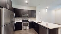 This single-floor residence showcases a contemporary design with a cohesive color palette of dark espresso tones and crisp white surfaces throughout. The kitchen serves as a central highlight, featuring rich dark cabinetry extending to the ceiling for ample storage, white quartz countertops, and a full suite of stainless steel appliances, including a refrigerator, electric range, built-in microwave, and dishwasher. A textured stone tile backsplash adds visual depth, while elegant pendant lighting illuminates the sink and peninsular counter area. Wood-look flooring runs throughout the kitchen and living areas, lending warmth and continuity to the open layout. The living area benefits from natural light through large windows and is equipped with a ceiling fan. French doors provide additional access and outdoor views. The bathroom maintains the same refined aesthetic with dark cabinetry, a white undermount sink, a large framed mirror, and light tile flooring, with an adjacent walk-in closet offering generous storage. The carpeted bedroom is spacious, bathed in natural light from a wide window, and includes a ceiling fan for comfort. Overall, the space presents a clean, modern aesthetic with thoughtful finishes.