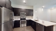 This single-floor residence showcases a contemporary design with a cohesive color palette of dark espresso tones and crisp white surfaces throughout. The kitchen serves as a central highlight, featuring rich dark cabinetry extending to the ceiling for ample storage, white quartz countertops, and a full suite of stainless steel appliances, including a refrigerator, electric range, built-in microwave, and dishwasher. A textured stone tile backsplash adds visual depth, while elegant pendant lighting illuminates the sink and peninsular counter area. Wood-look flooring runs throughout the kitchen and living areas, lending warmth and continuity to the open layout. The living area benefits from natural light through large windows and is equipped with a ceiling fan. French doors provide additional access and outdoor views. The bathroom maintains the same refined aesthetic with dark cabinetry, a white undermount sink, a large framed mirror, and light tile flooring, with an adjacent walk-in closet offering generous storage. The carpeted bedroom is spacious, bathed in natural light from a wide window, and includes a ceiling fan for comfort. Overall, the space presents a clean, modern aesthetic with thoughtful finishes.