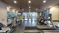 This fitness center offers a comprehensive workout environment thoughtfully designed to accommodate a variety of exercise routines. The space is anchored by floor-to-ceiling windows along the far wall, flooding the interior with natural light and providing open views of the surrounding exterior. Recessed ceiling lighting, complemented by multiple ceiling fans, ensures the space remains well-lit and well-ventilated throughout.

The layout is organized to separate strength training and cardio zones effectively. The strength training area features a multi-station cable machine, a weight bench, and a selection of resistance equipment, while an American flag-themed accent wall adds a distinctive visual element. Mounted televisions provide entertainment for users during their workouts.

The cardio section is lined with a variety of machines, including treadmills, ellipticals, and stationary bikes, all positioned to take advantage of the natural light from the large windows. The flooring transitions between rubberized matting in the free weights area and a smooth, durable surface throughout, supporting both safety and functionality. Overall, the space presents a clean, modern aesthetic well-suited for daily fitness use.