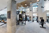 Located at 4015 South Blvd in Charlotte, NC, this professionally equipped fitness center offers a generously sized, single-floor layout divided into two distinct workout zones. The main fitness area impresses with soaring double-height ceilings, floor-to-ceiling windows that flood the space with natural light, and views of the surrounding building exterior. A textured geometric ceiling with pendant lighting adds an industrial-modern aesthetic, complemented by exposed concrete columns and neutral-toned carpet flooring throughout. This primary zone is outfitted with a comprehensive range of equipment, including cable and multi-station strength machines, treadmills, ellipticals, rowing machines, free weights, and functional training rigs. Full-length mirrors line the walls, enhancing the sense of openness. A secondary, more intimate room features a dedicated cycling studio with stationary bikes, warm wood built-in shelving, ceiling fans, and wall-to-wall mirrors, creating a bright and focused environment. An additional transitional area provides supplemental free weight storage, dumbbell racks, and core training equipment. The overall ambiance is modern, clean, and purposefully designed to support a wide variety of fitness activities in a well-lit, spacious setting.