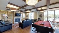 This amenity-rich space is designed to offer residents a variety of social and recreational experiences across multiple distinct areas. The interior game lounge is a standout, showcasing a coffered ceiling with rich wood beam detailing and statement drum pendant lighting. A stone-surround fireplace anchors one wall beneath a wall-mounted television, while hardwood floors, a leather seating arrangement, and a full-size pool table create an inviting atmosphere for both relaxation and entertainment. Large windows flood the room with natural light and offer views of the surrounding area. The outdoor courtyard is thoughtfully designed with stone pillar accents, a pergola overhead structure, and multiple seating clusters with fire pit tables — ideal for casual gatherings in a sheltered setting. The building's stone and wood exterior facade frames the space, lending a warm, lodge-inspired aesthetic. A rooftop terrace rounds out the amenities, featuring open-air dining areas, built-in grilling stations, and a decorative stone water feature wall as a central focal point, all set against open sky views of the surrounding urban landscape.