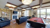 This amenity-rich space is designed to offer residents a variety of social and recreational experiences across multiple distinct areas. The interior game lounge is a standout, showcasing a coffered ceiling with rich wood beam detailing and statement drum pendant lighting. A stone-surround fireplace anchors one wall beneath a wall-mounted television, while hardwood floors, a leather seating arrangement, and a full-size pool table create an inviting atmosphere for both relaxation and entertainment. Large windows flood the room with natural light and offer views of the surrounding area. The outdoor courtyard is thoughtfully designed with stone pillar accents, a pergola overhead structure, and multiple seating clusters with fire pit tables — ideal for casual gatherings in a sheltered setting. The building's stone and wood exterior facade frames the space, lending a warm, lodge-inspired aesthetic. A rooftop terrace rounds out the amenities, featuring open-air dining areas, built-in grilling stations, and a decorative stone water feature wall as a central focal point, all set against open sky views of the surrounding urban landscape.