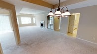 This single-floor space is designed with an open-concept layout that encourages a natural flow between the living, dining, and kitchen areas. The expansive carpeted living area provides a comfortable and versatile foundation, finished in soft neutral tones that complement the overall aesthetic of the space. A classic fireplace serves as a focal point in the living area, adding both warmth and character to the room.

A bronze-finished chandelier with globe shades hangs prominently, defining the dining area and providing ambient lighting. The ceiling features subtle architectural detailing, including a tray-style soffit accent highlighted in warm gold tones, adding visual interest and dimension to the space.

Adjacent to the living and dining areas, the kitchen is partially visible and features white cabinetry and light tile flooring, creating a clean, bright contrast to the carpeted areas. Built-in shelving is also visible nearby, offering practical storage solutions.

Large windows allow natural light to filter into the living area, brightening the neutral palette throughout. The cohesive design, open sightlines, and consistent use of warm, understated finishes give this space a welcoming and functional ambiance.