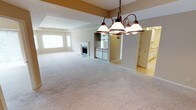 This single-floor space is designed with an open-concept layout that encourages a natural flow between the living, dining, and kitchen areas. The expansive carpeted living area provides a comfortable and versatile foundation, finished in soft neutral tones that complement the overall aesthetic of the space. A classic fireplace serves as a focal point in the living area, adding both warmth and character to the room.

A bronze-finished chandelier with globe shades hangs prominently, defining the dining area and providing ambient lighting. The ceiling features subtle architectural detailing, including a tray-style soffit accent highlighted in warm gold tones, adding visual interest and dimension to the space.

Adjacent to the living and dining areas, the kitchen is partially visible and features white cabinetry and light tile flooring, creating a clean, bright contrast to the carpeted areas. Built-in shelving is also visible nearby, offering practical storage solutions.

Large windows allow natural light to filter into the living area, brightening the neutral palette throughout. The cohesive design, open sightlines, and consistent use of warm, understated finishes give this space a welcoming and functional ambiance.