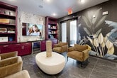 This single-floor amenity lounge is a thoughtfully designed social space that blends bold aesthetics with practical functionality. The most striking design element is the immersive large-scale botanical mural that spans multiple walls, depicting tropical foliage in muted greens, golds, and creams against a dark charcoal background, creating a dramatic and cohesive visual identity throughout the room.

The entertainment and lounge area features a built-in wet bar with deep plum-toned cabinetry, open shelving stocked with games and décor, a mounted large-screen television, and an integrated beverage cooler — ideal for hosting gatherings. A sculptural oval coffee table in a stone finish anchors a cluster of plush tan armchairs, encouraging relaxed social interaction. Complementing this is a contrasting floral-patterned accent wall that adds texture and warmth to the entertainment corner.

Toward the interior, a round dining table surrounded by velvet mustard-yellow chairs with gold-toned legs provides a dedicated space for more structured seating or gaming. The flooring throughout is a polished dark marble-look tile that ties the entire space together with sophistication. Recessed lighting and a decorative starburst pendant fixture illuminate the room evenly. A glazed entry door allows visual connectivity to adjacent spaces, rounding out this well-appointed communal lounge.