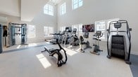 Located at 3420 Legacy Eagle Dr in Mount Pleasant, South Carolina, this modern fitness center is designed to support a comprehensive workout experience within a bright, thoughtfully organized single-floor layout. The facility is defined by soaring double-height ceilings and an abundance of natural light pouring in through large windows on multiple walls, creating an open and energizing atmosphere throughout.

The cardio zone is generously proportioned, housing a full lineup of equipment including treadmills, ellipticals, stationary bikes, and recumbent cycles, all positioned beneath the dramatic vaulted ceiling with wall-mounted televisions for entertainment. The strength training area features free weights, a dumbbell rack, a multi-station weight machine, and a bench press setup, complemented by floor-to-ceiling mirrors that visually expand the space.

A sleek glass partition with decorative vertical rods elegantly separates the main workout floor from a dedicated studio room, which is enclosed with glass walls and a sliding glass door. This studio is outfitted for group fitness or individual training, featuring a wall-mounted screen and open floor space. A stylish staircase with steel cable railings and wood-paneled accent walls adds an architectural focal point near the entrance corridor. Accent walls in bold teal, recessed lighting, and durable rubberized flooring throughout complete the polished, modern aesthetic of this well-appointed fitness facility.