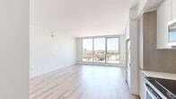 Located at 777 E Ocean Blvd in Long Beach, California, this thoughtfully designed unit offers a clean, modern aesthetic across a compact yet efficient single-floor layout comprising a living room, kitchen, bathroom, and hallway. The living area is the centerpiece of the space, highlighted by expansive floor-to-ceiling windows that flood the room with natural light and frame impressive panoramic views of the Long Beach cityscape, including palm tree-lined streets and neighboring buildings stretching toward the horizon. Light-toned hardwood flooring runs throughout the main areas, contributing to the bright and airy atmosphere. The bedroom area features built-in closet storage and continues the neutral, minimalist palette with crisp white walls. The kitchen is outfitted with stainless steel appliances, including a range and over-the-range microwave, complemented by white flat-panel cabinetry, light quartz countertops, and a textured white tile backsplash that adds subtle visual interest. The bathroom offers a functional and contemporary design with dark marble-tiled walls in the shower, built-in shelving, and dark wood-toned flooring that contrasts the lighter finishes found elsewhere in the unit. Overall, the space presents a clean, modern living environment with standout natural light and city views.