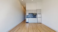 This single-floor unit showcases a contemporary open-concept design with impressively tall ceilings that amplify the sense of space throughout the main living area. Light wood-toned flooring runs continuously underfoot, lending warmth and cohesion to the clean, minimalist aesthetic defined by bright white walls and recessed lighting.

The kitchen is efficiently laid out along one wall, featuring flat-front white cabinetry extending to ceiling height for generous storage. A bold blue hexagonal tile backsplash provides a striking visual accent against the neutral palette. Stainless steel appliances — including a refrigerator, microwave, and dishwasher — complete the functional setup.

The living area is generously proportioned and receives abundant natural light through a large sliding glass door, which opens to an exterior view of the surrounding neighborhood and fills the space with sunlight.

The bathroom is thoughtfully designed, incorporating a glass-enclosed shower with vertically laid tile, a toilet, and a dedicated in-unit washer, all finished with the same light wood flooring for a unified feel. Chrome fixtures and a built-in shelving unit add practical elegance to the space.