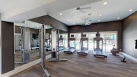 Located at 901 Time Centre Dr in Saint Charles, Missouri, this fitness center is a modern, thoughtfully designed facility built to support a full range of workout routines. The space is finished with wood-look flooring throughout and features bold dark gray walls accented by crisp white trim, creating a sleek and motivating atmosphere. Recessed lighting and ceiling fans keep the environment well-lit and comfortable during workouts.

The main exercise floor is generously sized and houses a row of treadmills equipped with individual screens, a cable strength training machine, elliptical trainers, stationary bikes, a rowing machine, and a dedicated dumbbell rack with a full selection of free weights. Adjustable benches provide additional options for strength-focused training. Floor-to-ceiling mirrors line one wall, enhancing the sense of space and allowing users to monitor their form.

Large windows and sliding glass doors allow ample natural light to flow into the space while offering views of the outdoor pool area, creating an inviting connection between indoor exercise and the surrounding amenity spaces. A separate studio room with frosted glass panel doors provides a quieter area for stretching, yoga, or mat-based workouts, complete with exercise balls and yoga mats. An entry corridor with wood-look flooring connects the main entrance to the various workout areas, completing this well-appointed fitness facility.