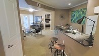 This single-floor residence showcases an open-concept design that effortlessly connects the main living areas. The kitchen features a granite-topped breakfast bar with rustic wooden bar stools, creating a casual dining spot that overlooks the rest of the living space. Adjacent to the bar, a dedicated dining area accommodates a small table with teal accent chairs, adding a pop of color to the neutral palette. The living room is finished with plush carpeting and is bathed in natural light streaming through large windows dressed with blinds. A ceiling fan keeps the space comfortable, and crown molding throughout lends a polished, refined touch to the interior. The warm taupe wall tones create a cohesive and inviting ambiance across the open floor plan. Decorative wall art and a sunburst mirror accent the walls, adding character and visual interest. A glimpse through an open doorway reveals a well-appointed bedroom, seamlessly integrated into the overall layout. The combination of neutral tones, modern furnishings, and thoughtful design details makes this a comfortable and stylish single-level living space.