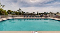 Located in the Ashland Cir community of Fort Pierce, Florida, this outdoor pool facility offers a well-appointed recreational space designed for relaxation and leisure. The centerpiece is a large, rectangular swimming pool with vivid turquoise water and clearly marked depth indicators, providing a safe and inviting environment for swimmers of all ages. Adjacent to the main pool is a smaller wading pool, ideal for younger visitors or those seeking a more relaxed experience.

The expansive paver deck surrounds the pool on all sides, offering ample space for lounging. Numerous blue sling-style lounge chairs and upright chairs are arranged throughout the deck, accompanied by umbrella-shaded seating areas for relief from the Florida sun. A covered pavilion with a pitched roof and decorative columns is positioned along one side of the pool area, providing a shaded gathering space with ceiling fans for additional comfort.

The facility is fully enclosed by a white metal perimeter fence, ensuring safety and privacy. Lush native landscaping, including tall palms and mature pine trees, frames the space and creates a natural, tranquil backdrop. A children's play structure is also visible within the grounds. The overall design reflects a clean, modern Florida aesthetic, making this community pool area a functional and attractive amenity.