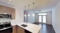 This single-floor residence showcases a thoughtfully designed open-concept layout that seamlessly connects the kitchen and living areas. The kitchen serves as a central hub, anchored by a spacious island with a crisp white quartz countertop and an undermount stainless steel sink with a sleek arc faucet. Warm wood-toned cabinetry with brushed hardware provides generous storage and introduces an organic contrast to the bright, clean surfaces. A built-in appliance, likely a dishwasher, is integrated beneath the island for a streamlined look.

Three cylindrical pendant lights hang at varying heights above the island, providing focused task lighting while contributing a modern aesthetic. Dark hardwood-style flooring runs throughout, grounding the space with a rich, durable surface. The adjacent living area benefits from abundant natural light streaming through double French doors fitted with horizontal blinds, offering both brightness and privacy. A ceiling fan is positioned overhead in the living space, ensuring year-round comfort. The overall ambiance is clean, contemporary, and functional, with a neutral color palette that creates a welcoming and versatile living environment.