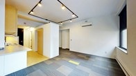 This single-floor space presents a clean, modern aesthetic defined by thoughtful design details and practical finishes throughout. The main open area is anchored by striking black track lighting that runs across the ceiling, providing both functional illumination and a bold industrial character. The flooring features multi-toned modular carpet tiles arranged in a geometric pattern, lending texture and visual interest to the space, with a contrasting yellow accent tile adding a subtle pop of color. Crisp white walls and a smooth ceiling create a bright, neutral backdrop that enhances the sense of openness. A wall-mounted split air conditioning unit ensures year-round climate comfort. A partial partition wall divides the interior, creating a sense of zoning without fully enclosing separate areas, allowing the space to maintain an open and connected feel. Beyond the partition, a secondary area is accessible, offering additional flexible space. To one side, a transition to a lighter wood-toned floor leads toward what appears to be a utility or secondary access area with a wooden door, adding a warm contrast to the otherwise contemporary palette. Overall, the space communicates a versatile, well-lit environment suited to a variety of uses, combining modern design elements with a functional and adaptable layout.