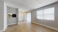 This single-floor unit offers a practical and clean layout designed for comfortable everyday living. The main living area is generously proportioned, featuring light wood-look flooring that runs consistently throughout the space, neutral grey walls, and a textured popcorn ceiling. Multiple windows fitted with horizontal blinds allow natural light to filter in, while a sliding glass door with vertical blinds provides access to an outdoor area. Two louvered closet doors in the living zone offer ample built-in storage.

The galley-style kitchen is efficiently arranged, featuring white shaker-style cabinetry with brushed nickel hardware, light granite-look countertops, and black appliances including an electric range and refrigerator. A pass-through opening connects the kitchen to the living area, enhancing the sense of openness. A tile backsplash accent adds a subtle decorative touch.

A hallway leads to the bathroom, which includes a full-size soaking tub and shower combination enclosed by classic white square wall tiles. The wood-look flooring continues into the hallway, maintaining visual continuity throughout. An electrical panel is conveniently accessible from the hallway. The overall ambiance is bright, clean, and functional.