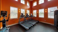 Located at 810 Eden Ave in Fort Mill, SC, this single-floor fitness center is designed to energize and motivate. The space is immediately distinguished by its bold, vibrant orange walls that create a dynamic and high-energy atmosphere throughout. Durable rubber flooring covers the entire area, providing a safe and functional surface suited for a wide range of workout activities.

The cardio zone features a row of treadmills positioned beneath soaring double-height ceilings, flanked by large windows on multiple levels that flood the area with natural light and offer views of the surrounding outdoors. Wall sconces add warm accent lighting to complement the bright, open feel.

A dedicated strength training area houses a multi-functional cable machine, a weight rack stocked with medicine balls and free weights, and a pull-up bar mounted to the wall. A suspended heavy punching bag adds a boxing-focused training option. An additional area accommodates a rowing machine, an elliptical trainer, and a stationary bike, offering versatile low-impact cardio options.

Ceiling fans are mounted throughout to maintain air circulation during intense workouts. A large mirror is strategically placed to allow users to monitor their form. Recessed lighting and natural light from multiple windows ensure the space remains bright and welcoming, making this fitness center a well-equipped and thoughtfully designed facility.
