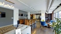 This single-floor space presents a sleek, contemporary design that blends functionality with modern aesthetics. The open-plan layout seamlessly connects multiple zones, creating a dynamic and inviting environment.

At the heart of the space is a prominent bar-height counter crafted with warm wood cabinetry, paired with a row of chrome and wood swivel bar stools. Overhead, a striking cluster of pendant globe lights hangs at varying heights, adding a sculptural focal point to the area. Adjacent to the counter, a dark textured tile backsplash serves as a bold accent wall, complemented by wall-mounted flat-screen televisions, suggesting a media or entertainment function.

The lounge area is furnished with a plush, light grey sofa and a pair of stylish accent chairs in blue and green tones, arranged to encourage conversation and relaxation. Dark polished concrete floors run throughout, grounding the space with an industrial yet refined character.

Exposed structural columns and a ceiling fitted with track lighting provide both architectural interest and practical illumination. The open sightlines extend deeper into the space, hinting at additional areas beyond. The overall ambiance is modern, professional, and welcoming — well-suited for a hospitality, co-working, or residential amenity environment.