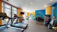 This fitness center offers a well-equipped, energetic workout environment spread across a single floor. The space is visually defined by bold orange and teal-blue accent walls, recessed ceiling lighting, and rubber flooring throughout, creating a lively yet functional atmosphere. Architectural details include a decorative column and large windows along one wall that invite natural light and views of the surrounding building exterior.

The cardio zone features a row of True-brand treadmills and elliptical machines aligned along the mirrored wall, maximizing the sense of space. A spin bike, air bike, and rowing machine occupy a separate area alongside wall-mounted flat-screen televisions for entertainment during workouts. Stability and medicine balls are neatly stored nearby for functional training use.

The strength training section includes a multi-station cable machine, a dedicated dumbbell rack ranging across multiple weight increments, kettlebells, and a weight bench, offering a comprehensive range of resistance training options. Motivational posters add a spirited touch to the teal accent wall in this zone.

A mirrored glass partition separates a secondary area, adding depth to the layout. Overall, the fitness center balances a compact, efficient footprint with a diverse array of equipment suited for cardio, strength, and functional training.