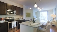 This well-appointed single-floor residence offers a thoughtfully designed open-concept layout that seamlessly connects the kitchen, dining, and living areas. The kitchen is a standout feature, showcasing rich dark espresso cabinetry paired with light granite countertops that extend into a curved breakfast bar — ideal for casual dining or entertaining. Stainless steel appliances, including a range, microwave, and refrigerator, provide a sleek, modern aesthetic. Elegant pendant lighting hangs above the kitchen island, adding warmth and a refined touch to the space. The open flow from the kitchen leads naturally into the living and dining areas, where large windows and a sliding glass door flood the interior with natural light, creating a bright and airy ambiance throughout. The neutral color palette, featuring soft white and warm gray tones, complements the hardwood-style flooring and contributes to a clean, cohesive look. Decorative wall art and thoughtful furnishings complete the inviting atmosphere, making this residence both functional and visually appealing. The generous natural light and open sightlines give the space a sense of openness and ease.