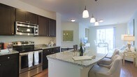 This well-appointed single-floor residence offers a thoughtfully designed open-concept layout that seamlessly connects the kitchen, dining, and living areas. The kitchen is a standout feature, showcasing rich dark espresso cabinetry paired with light granite countertops that extend into a curved breakfast bar — ideal for casual dining or entertaining. Stainless steel appliances, including a range, microwave, and refrigerator, provide a sleek, modern aesthetic. Elegant pendant lighting hangs above the kitchen island, adding warmth and a refined touch to the space. The open flow from the kitchen leads naturally into the living and dining areas, where large windows and a sliding glass door flood the interior with natural light, creating a bright and airy ambiance throughout. The neutral color palette, featuring soft white and warm gray tones, complements the hardwood-style flooring and contributes to a clean, cohesive look. Decorative wall art and thoughtful furnishings complete the inviting atmosphere, making this residence both functional and visually appealing. The generous natural light and open sightlines give the space a sense of openness and ease.