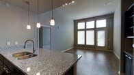 This single-floor space showcases a thoughtfully designed open layout that seamlessly connects the kitchen and living areas. The kitchen is anchored by a generous granite-topped island with a built-in sink, offering both functional workspace and casual seating. Pendant lights hang above the island, complementing the track lighting installed along the ceiling to provide layered illumination throughout.

The soaring ceilings lend a sense of openness and grandeur, enhancing the airy atmosphere of the interior. Rich dark hardwood flooring flows consistently through the living area, adding warmth and a polished aesthetic. The living space is bathed in natural light courtesy of a large, multi-pane window arrangement paired with a glass-paneled door, both fitted with horizontal blinds for adjustable privacy.

Neutral gray-toned walls provide a clean, contemporary backdrop that complements the dark flooring and granite finishes. A hallway visible from the kitchen hints at additional rooms beyond, offering a sense of depth within the floor plan. Overall, the space strikes a balance between modern sophistication and practical, livable design.