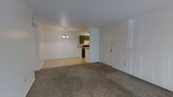 This single-floor residence features an open-concept layout that combines the living and dining areas into one cohesive space. The living area is finished with wall-to-wall carpet in a neutral tone, providing a comfortable underfoot feel, while the adjoining dining area transitions to hard flooring. A modest chandelier hangs above the dining space, adding a subtle decorative touch to the otherwise straightforward design. The walls are painted in a clean white throughout, contributing to a bright and airy atmosphere.

Visible from the main living area, the kitchen is partially open to the dining space via a pass-through counter, allowing for convenient interaction between the two areas. The kitchen features dark wood cabinetry and appears compact yet functional, with appliances integrated beneath the upper cabinets. A range hood is visible above the cooking area.

A solid exterior door is positioned along one wall, providing direct access to the outside. The layout is practical and efficient, with a clear flow between the main living spaces. The overall design is straightforward and functional, suited for everyday living with a neutral palette that offers flexibility for a variety of interior styles.