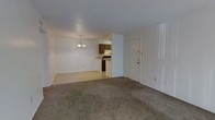 This single-floor residence features an open-concept layout that combines the living and dining areas into one cohesive space. The living area is finished with wall-to-wall carpet in a neutral tone, providing a comfortable underfoot feel, while the adjoining dining area transitions to hard flooring. A modest chandelier hangs above the dining space, adding a subtle decorative touch to the otherwise straightforward design. The walls are painted in a clean white throughout, contributing to a bright and airy atmosphere.

Visible from the main living area, the kitchen is partially open to the dining space via a pass-through counter, allowing for convenient interaction between the two areas. The kitchen features dark wood cabinetry and appears compact yet functional, with appliances integrated beneath the upper cabinets. A range hood is visible above the cooking area.

A solid exterior door is positioned along one wall, providing direct access to the outside. The layout is practical and efficient, with a clear flow between the main living spaces. The overall design is straightforward and functional, suited for everyday living with a neutral palette that offers flexibility for a variety of interior styles.
