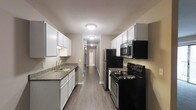 This single-floor residential space is designed with a functional and clean aesthetic that makes efficient use of its layout. The galley-style kitchen serves as a central highlight, featuring white shaker-style cabinetry that lines both walls, providing ample storage space. Dark granite countertops offer a striking contrast against the white cabinets, while a stainless steel sink with a modern faucet adds a practical touch. Black appliances — including a refrigerator, electric range, over-the-range microwave, and dishwasher — create a cohesive and contemporary look throughout the kitchen. Warm-toned wood-look flooring runs continuously from the kitchen down a hallway that connects to the rest of the unit, lending a sense of flow and continuity to the space. Flush-mount ceiling light fixtures provide consistent overhead illumination throughout. The neutral wall tones in warm beige complement the overall color palette, creating a welcoming and versatile atmosphere. The corridor leading away from the kitchen hints at additional rooms beyond, suggesting a well-organized floor plan. Overall, the space balances practicality with modern design sensibilities, making it a comfortable and functional living environment.