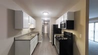 This single-floor residential space is designed with a functional and clean aesthetic that makes efficient use of its layout. The galley-style kitchen serves as a central highlight, featuring white shaker-style cabinetry that lines both walls, providing ample storage space. Dark granite countertops offer a striking contrast against the white cabinets, while a stainless steel sink with a modern faucet adds a practical touch. Black appliances — including a refrigerator, electric range, over-the-range microwave, and dishwasher — create a cohesive and contemporary look throughout the kitchen. Warm-toned wood-look flooring runs continuously from the kitchen down a hallway that connects to the rest of the unit, lending a sense of flow and continuity to the space. Flush-mount ceiling light fixtures provide consistent overhead illumination throughout. The neutral wall tones in warm beige complement the overall color palette, creating a welcoming and versatile atmosphere. The corridor leading away from the kitchen hints at additional rooms beyond, suggesting a well-organized floor plan. Overall, the space balances practicality with modern design sensibilities, making it a comfortable and functional living environment.