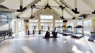 Located at 3530 Brookstone Dr in Cincinnati, OH, this single-story fitness center is architecturally distinguished by its dramatic vaulted ceiling with exposed white-painted beam trusses and dark accent supports, creating a striking, open atmosphere. Ceiling fans and track lighting run along the roofline, ensuring the space remains well-lit and comfortable throughout. Large windows wrap around multiple walls, flooding the interior with natural light while offering direct views of an outdoor pool and surrounding landscape, enhancing the workout environment with a bright, energizing ambiance.

The durable diamond-plate interlocking floor tiles cover the expansive main workout area, which is equipped with a well-rounded selection of cardio machines, including multiple treadmills, ellipticals, and a stationary bike. A dedicated strength-training zone features a dumbbell rack with a full weight selection, a wall-mounted television, and a bold black accent wall, providing a focused space for resistance training. An additional strength machine is positioned near the entry area.

The facility includes a glass-enclosed entry vestibule and a small interior room, likely for storage or administrative use. Yellow accent walls throughout add a vibrant, motivating touch to the overall design, making this a well-appointed and visually appealing fitness facility.