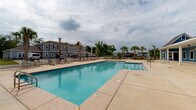 Located at 3867 Stardust Dr in Foley, Alabama, this outdoor amenity space centers around a large, clear-water swimming pool that serves as the focal point of a well-appointed community recreation area. The pool is rectangular in design with a shallow wading section, framed by an expansive concrete sundeck that provides ample room for lounging and socializing.

A covered pavilion runs along one side of the pool area, offering a shaded retreat with ceiling fans and direct access to adjacent facilities — ideal for relaxing out of the sun. The deck is furnished with lounge chairs and umbrella-shaded seating arrangements, creating a comfortable outdoor gathering space.

The pool area is enclosed by a black metal fence, providing both security and a clean visual boundary. Tall palm trees are planted throughout the surrounding grounds, lending a coastal, resort-like ambiance that complements the warm Alabama climate. The multi-building residential community, featuring blue-gray exterior siding and pitched brown rooflines, is visible in the background, reinforcing the communal nature of the space.

Overall, this outdoor pool area is thoughtfully designed to deliver a relaxing, resort-style experience, combining practical amenities with an inviting atmosphere well-suited to the Gulf Coast region.