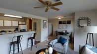 This single-floor residence offers an open-concept layout that seamlessly connects the kitchen, dining, and living areas into one cohesive, functional space. The living room is carpeted and furnished with a patterned armchair accented by a navy blue throw pillow, centered around a dark circular coffee table. A ceiling fan with wooden blades serves as a practical overhead fixture, complementing the standard ceiling height throughout. Framed wall art and a decorative vase with branches add character to the space.

The dining area sits adjacent to the living room, featuring a round dark wood table surrounded by traditional-style chairs, positioned beneath a flush-mount light fixture. A trio of abstract wall panels adds a subtle artistic touch nearby.

The kitchen is visible through a pass-through opening, highlighted by a light-colored breakfast bar with tall industrial-style bar stools. Cabinetry and basic appliances, including a refrigerator, are visible in the background. The kitchen features wood-toned flooring, contrasting with the carpeted living and dining areas. A doorway visible in the background leads to additional rooms, suggesting a practical and well-organized floor plan throughout.