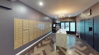 This single-floor common area serves as a multifunctional lobby space combining mail retrieval, package pickup, and a convenient market. The space is finished with a cohesive design palette of deep gray-purple walls, crisp white baseboards, and rich herringbone-patterned wood flooring that runs throughout, lending the area a sophisticated, contemporary feel.

An extensive wall of gold-finished individual mailboxes lines one side of the space, numbered to serve a large number of residents and accompanied by several oversized parcel lockers for package deliveries. Opposite and adjacent to the mailboxes, the Tribeca Market area offers a curated retail experience, featuring open shelving stocked with snack items under a "Shop" sign, alongside illuminated glass-door refrigerators displaying a wide selection of beverages and chilled goods under the "Market" designation.

A central island with a white marble-style countertop and gray cabinetry accented with gold hardware provides a functional workspace within the market area. Above it, a gold multi-arm chandelier adds an upscale touch. A wall-mounted flat-screen television is present in an adjacent open area, suggesting a lounge or waiting zone. Recessed lighting throughout ensures the entire space is well-illuminated and welcoming.