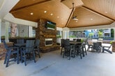 Located at 1570 Sheridan Rd NE in Atlanta's vibrant 30324 corridor, this outdoor amenity space offers a thoughtfully designed communal environment for relaxation and social gathering. At the heart of the space is a striking covered pavilion featuring a soaring octagonal wood-paneled ceiling with recessed lighting and a large ceiling fan, providing comfort in both warm and cool seasons. A prominent stacked-stone fireplace anchors the pavilion's interior wall and is topped with a mounted flat-screen television, making it an ideal setting for outdoor entertaining. Multiple high-top and standard dining tables with contemporary chairs provide ample seating throughout the covered area. Stone-clad pillar accents lend architectural character to the pavilion's open-air perimeter, which flows seamlessly onto a broad concrete patio. Beyond the covered structure, the space opens to a well-manicured lawn framed by young trees, garden beds with shrubs, and wooden bench seating. An outdoor grilling station is conveniently positioned nearby for al fresco dining. The surrounding residential buildings, finished in neutral and blue-toned siding with balconies, frame the courtyard and contribute to an intimate neighborhood atmosphere. Lush greenery and mature trees further enhance the tranquil, park-like setting.