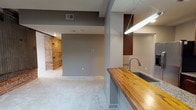 This single-floor space masterfully combines industrial heritage with contemporary design. Exposed red brick walls serve as a striking architectural focal point throughout the living areas, complemented by a rustic sliding barn door that adds both character and function. Multiple skylights flood the space with natural light, while ceiling fans ensure year-round comfort.

The kitchen is well-appointed with rich dark wood cabinetry, granite countertops, a full suite of stainless steel appliances, and a warm butcher block island counter that doubles as a casual dining surface. Track lighting illuminates the kitchen and adjacent open living area, which features plush neutral carpet underfoot.

The bedroom is a generous, light-filled retreat showcasing exposed dark wood structural beams that add warmth and a loft-like aesthetic. A skylight overhead and a ceiling fan enhance the room's open, airy feel, and a walk-in closet provides ample storage.

The bathrooms are finished with clean, modern tile work. One features a walk-in shower with white subway tile surround and chrome fixtures, while the other includes a granite-topped vanity with an undermount sink and matching dark wood cabinetry. Throughout the space, neutral gray walls tie together the blend of raw, industrial elements and refined modern details.