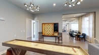 This well-appointed game room is designed to be both functional and inviting, offering a variety of entertainment options in a polished, contemporary setting. The room features warm hardwood-style flooring that flows seamlessly throughout, complemented by soft gray walls that create a relaxed yet refined atmosphere. Statement modern chandeliers with metallic cylindrical shades provide ample overhead illumination, lending the space a sophisticated touch.

A full-length shuffleboard table takes center stage in the foreground, crafted with a rich wood surround and a smooth playing surface — an ideal centerpiece for friendly competition. Against the wall, an oversized Scrabble board mounted on a wooden frame is paired with two bar-height chairs, creating a cozy nook for word game enthusiasts. A chalkboard panel adjacent to the Scrabble display adds a practical and charming element for scorekeeping or messaging.

The space opens through a wide passage into an adjacent lounge and dining area, where a large table surrounded by upholstered chairs and generous natural light from expansive windows can be seen, extending the social possibilities of the environment. A frosted glass door leads to an adjoining room, adding privacy while maintaining an open, airy feel throughout.
