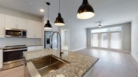 This single-story home in Dripping Springs, Texas offers a thoughtfully designed open-concept layout that seamlessly connects the kitchen, dining, and living areas. The kitchen serves as a central focal point, showcasing a large granite-topped island with an undermount stainless steel sink and a sleek pull-down faucet. Three matte black pendant lights hang above the island, complementing white shaker cabinetry, a subtle tile backsplash, stainless steel appliances, and a built-in beverage refrigerator. Light wood-look flooring flows throughout the main living areas, enhancing the bright, airy atmosphere.

The living room features a ceiling fan and French doors with transom windows that invite natural light and provide outdoor access. Bedrooms are carpeted for comfort, each equipped with ceiling fans and windows fitted with blinds. The primary bathroom includes a granite-topped double vanity, a glass-enclosed walk-in shower, and large-format tile flooring. A secondary bathroom mirrors the refined aesthetic with a granite vanity and a tiled tub-shower combination. Clean, neutral tones and modern finishes throughout lend the home a cohesive, contemporary character.