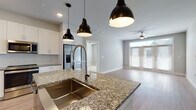 This single-story home in Dripping Springs, Texas offers a thoughtfully designed open-concept layout that seamlessly connects the kitchen, dining, and living areas. The kitchen serves as a central focal point, showcasing a large granite-topped island with an undermount stainless steel sink and a sleek pull-down faucet. Three matte black pendant lights hang above the island, complementing white shaker cabinetry, a subtle tile backsplash, stainless steel appliances, and a built-in beverage refrigerator. Light wood-look flooring flows throughout the main living areas, enhancing the bright, airy atmosphere.

The living room features a ceiling fan and French doors with transom windows that invite natural light and provide outdoor access. Bedrooms are carpeted for comfort, each equipped with ceiling fans and windows fitted with blinds. The primary bathroom includes a granite-topped double vanity, a glass-enclosed walk-in shower, and large-format tile flooring. A secondary bathroom mirrors the refined aesthetic with a granite vanity and a tiled tub-shower combination. Clean, neutral tones and modern finishes throughout lend the home a cohesive, contemporary character.