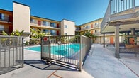 Located at 2700 Ellendale Place in Los Angeles, CA, this outdoor common area serves as the central hub of a multi-story residential property. The centerpiece of the space is a generously sized, resort-style swimming pool with striking turquoise water, enclosed by a sleek black metal safety fence with a gated entry. The pool area is surrounded by a spacious concrete deck outfitted with lounge chairs, providing ample room for relaxation under the Southern California sun. Lush tropical plantings, including mature banana trees, are thoughtfully positioned throughout the courtyard, adding vibrant greenery and a resort-like atmosphere. The surrounding building features multiple floors of residential units, each with private balconies that overlook the courtyard. The exterior facade is finished in warm earth tones with colorful accent doors in red and orange, contributing to the property's welcoming character. A covered ground-floor entry area offers a shaded seating nook with a decorative bench, providing a comfortable transition space between the building's interior and the outdoor courtyard. The open-air design of the courtyard maximizes natural light and takes full advantage of Los Angeles's year-round favorable climate, making this communal outdoor space a defining feature of the property.