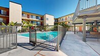 Located at 2700 Ellendale Place in Los Angeles, CA, this outdoor common area serves as the central hub of a multi-story residential property. The centerpiece of the space is a generously sized, resort-style swimming pool with striking turquoise water, enclosed by a sleek black metal safety fence with a gated entry. The pool area is surrounded by a spacious concrete deck outfitted with lounge chairs, providing ample room for relaxation under the Southern California sun. Lush tropical plantings, including mature banana trees, are thoughtfully positioned throughout the courtyard, adding vibrant greenery and a resort-like atmosphere. The surrounding building features multiple floors of residential units, each with private balconies that overlook the courtyard. The exterior facade is finished in warm earth tones with colorful accent doors in red and orange, contributing to the property's welcoming character. A covered ground-floor entry area offers a shaded seating nook with a decorative bench, providing a comfortable transition space between the building's interior and the outdoor courtyard. The open-air design of the courtyard maximizes natural light and takes full advantage of Los Angeles's year-round favorable climate, making this communal outdoor space a defining feature of the property.