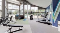 This single-floor fitness center is designed to deliver a bright, motivating workout environment. The space is anchored by dramatic vaulted ceilings with exposed wooden beams, soaring to impressive heights and creating an open, airy atmosphere throughout. Floor-to-ceiling window walls flood the interior with abundant natural light while offering views of lush surrounding greenery and an adjacent outdoor pool area, seamlessly connecting the indoors with the outdoors.

The flooring transitions between neutral carpet tiles and vibrant green turf zones, delineating dedicated areas for different training activities. The cardio section is well-appointed with treadmills, stationary bikes, and elliptical machines, while the strength training area features a multi-use cable machine, adjustable benches, and a full dumbbell rack. A functional training zone on the turf provides space for stretching, mat work, and step training, supported by a built-in storage unit stocked with yoga mats and foam rollers.

A bold, geometric accent wall in deep navy blue with geometric multi-color patterns adds a dynamic visual focal point to one corner of the space. Structural columns and a ceiling fan complement the architectural character of the room. The fitness center is accessible via a warm wood-framed glass door, and emergency exit signage ensures safe navigation throughout.
