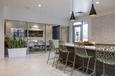 This thoughtfully designed single-floor communal space offers a seamless blend of social and relaxation areas within a clean, contemporary aesthetic. The entry foyer sets a welcoming tone with herringbone-patterned light tile flooring, tall potted plants flanking a console table, decorative artwork, and a diamond-pattern built-in bookshelf that transitions naturally into the main lounge.

The central lounge area provides generous seating with plush sofas, accent chairs, and coordinating ottomans arranged around a large area rug. A wall-mounted flat-screen TV anchors one end of the seating zone, while a billiards table positioned nearby adds a recreational focal point to the space.

The fully equipped kitchen features warm wood-grain cabinetry, white quartz countertops, mosaic tile backsplash, stainless steel appliances, and a spacious island with seating for several. Striking pendant lights hang above the island, and large windows allow abundant natural light while framing views of an adjacent outdoor patio area, which is accessible through a full-height glass door. A separate, glassed-in workspace or meeting room is also visible within the layout, rounding out the versatile functionality of this amenity space.