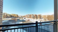 This single-floor residence offers a well-considered layout designed for comfortable, functional living. One of its most notable features is the private balcony, enclosed by a dark metal railing and supported by a sturdy post. The balcony provides expansive views of the surrounding brick multi-story residential community, open snow-covered grounds, mature bare-branched trees, and a clear blue sky — a serene winter panorama that brings a strong sense of openness to the unit.

Inside, the rooms are finished with neutral-toned carpet flooring and painted in soft, light gray and warm white hues, creating a calm and cohesive atmosphere throughout. Multiple windows fitted with horizontal blinds allow natural light to filter in from several directions, brightening each room. A corner seating area benefits from dual windows, maximizing light while offering pleasant outdoor sightlines.

One flexible room functions well as a home office or den, featuring a window nook that accommodates a writing desk. Adjacent to this space is a carpeted room with two built-in closets equipped with hanging rods and shelving, offering practical storage. The layout connects rooms smoothly, with clean, modern interior doors providing clear separation between spaces. Overall, the unit presents a tidy, light-filled environment with practical storage and appealing outdoor views.