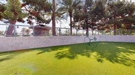 Located at 12375 W Van Buren St in Avondale, Arizona, this dedicated outdoor dog park offers a well-maintained, pet-friendly environment designed for both play and relaxation. The space is blanketed in vibrant artificial turf, providing a clean and comfortable surface for pets year-round in the Arizona climate. A covered pavilion with a modern flat roof and sleek steel posts anchors one corner of the area, offering shaded respite for owners and animals alike. The park is equipped with agility-style features, including a tunnel and a raised platform, encouraging active play. A paw-print-shaped agility ring adds a playful design element to the grounds. The space is fully enclosed by a combination of concrete block walls and metal fencing, ensuring a safe and secure environment. Waste disposal stations are conveniently positioned throughout the area. Mature palm trees and evergreen landscaping line the perimeter beyond the fencing, providing a lush visual backdrop and natural shade. The open layout allows for ample room to roam, while the surrounding multi-story residential building contextualizes this as a community amenity. Under the expansive Arizona sky, the park delivers a bright, open-air experience with functional design and thoughtful amenities.